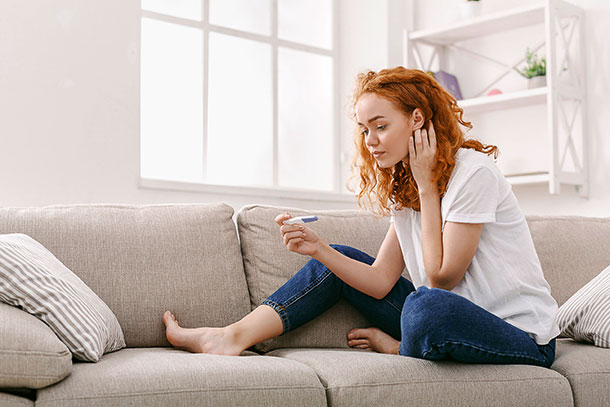 Eine junge Frau sitzt auf dem Sofa und betrachtet einen Schwangerschaftstest in ihrer Hand.
