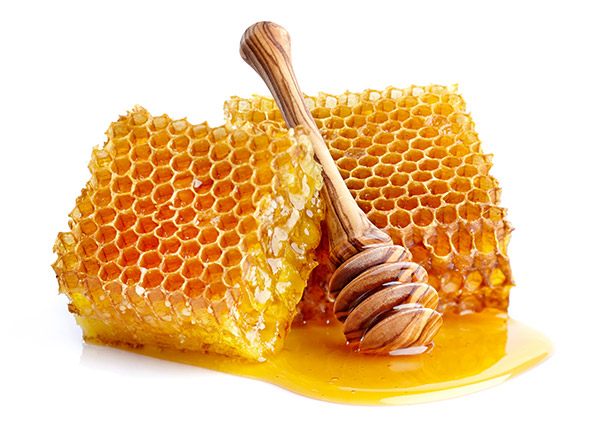 Zwei mit Honig triefende Bienenwaben liegen gut ersichtlich im weissen Raum. Ein hölzerner Honiglöffel liegt dabei.